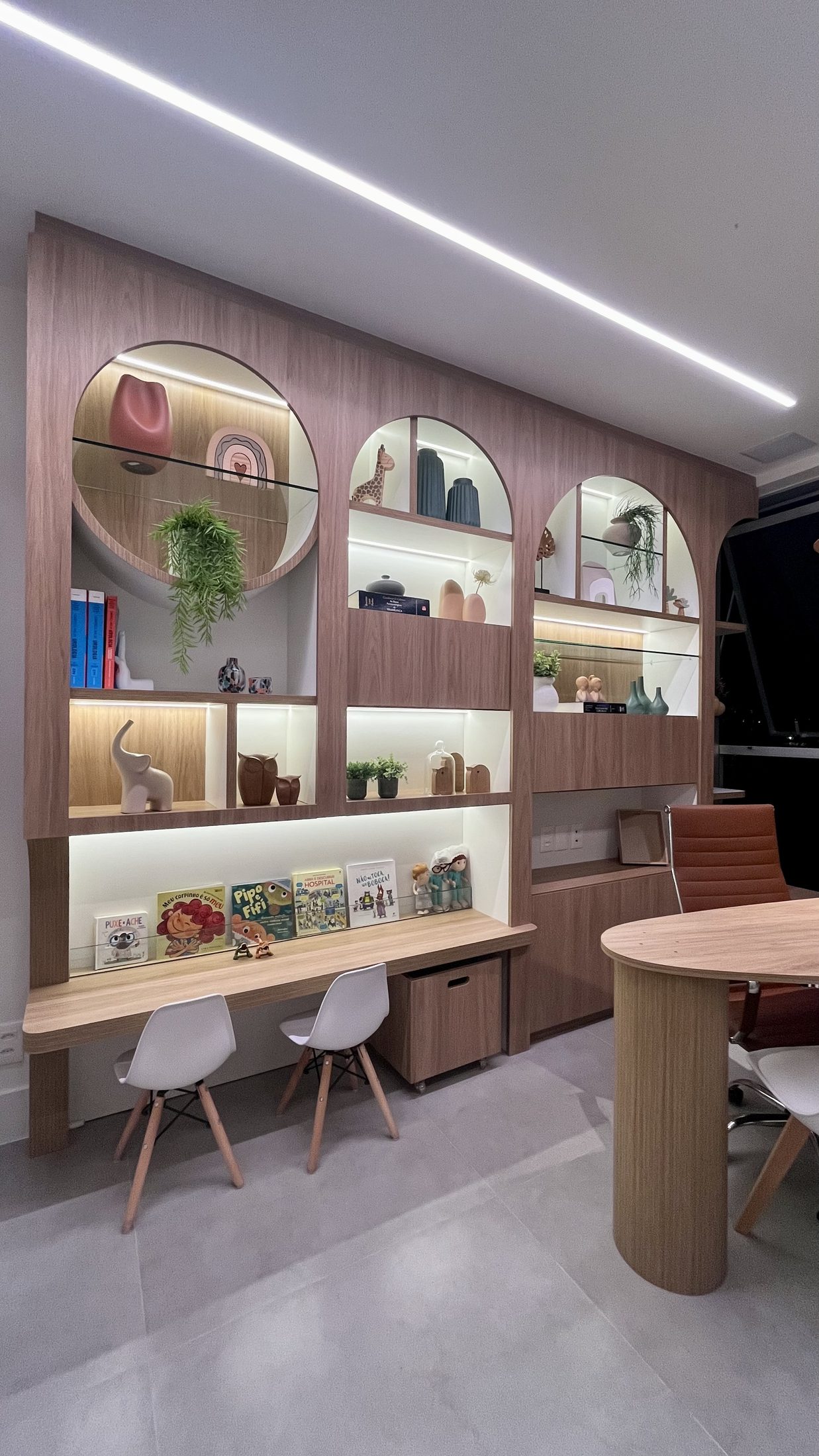 Sala de atendimento com marcenaria em arcos, bancada de madeira, livros infantis e cadeiras brancas